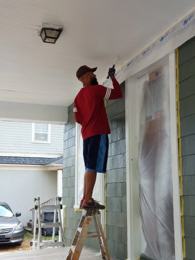 A painter working on the ceiling and trim of a taped and masked front porch