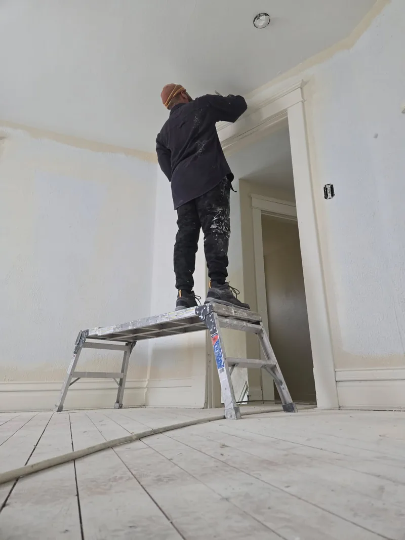 A painter on a platform ladder painting the ceiling edge and trim in a primed room